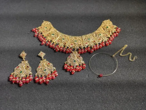Elegant gold-tone women's jewelry set, including a captivating step necklace, Maang Tika, Earrings and Nose Ring adorned with vibrant red beads for a touch of sophistication and style