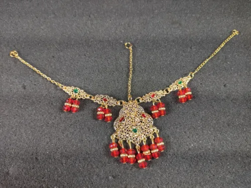 Elegant gold-tone women's jewelry set, including a captivating step necklace, Maang Tika, Earrings and Nose Ring adorned with vibrant red beads for a touch of sophistication and style