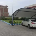 A car parked under a fabric car parking shade structure with advanced technology designed to provide effective sun protection while maintaining airflow. The PVC tensile fabric is stretched tautly over a sturdy frame, with sensors integrated for automated adjustments based on sunlight intensity and wind conditions.